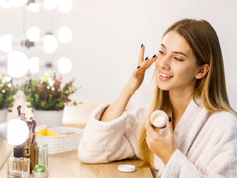 Young Beautiful Blonde Girl Cares Face Skin With Moisturizer Front Mirror