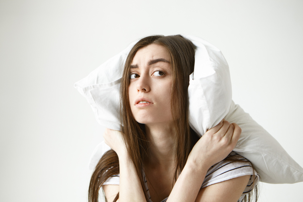 Young Brunette Woman With Messy Hairstyle Covering Ears Using White Pillow Looking Sideways With Frustration