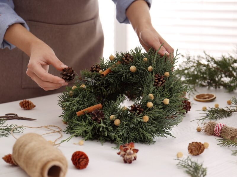 Florist Making Beautiful Christmas Wreath At White Wooden Table Indoors, Closeup