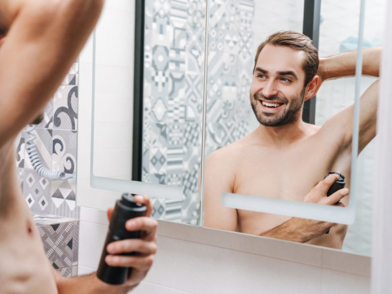 Smiling Young Shirtless Man Applying Deodorant While Standing Bathroom Mirror