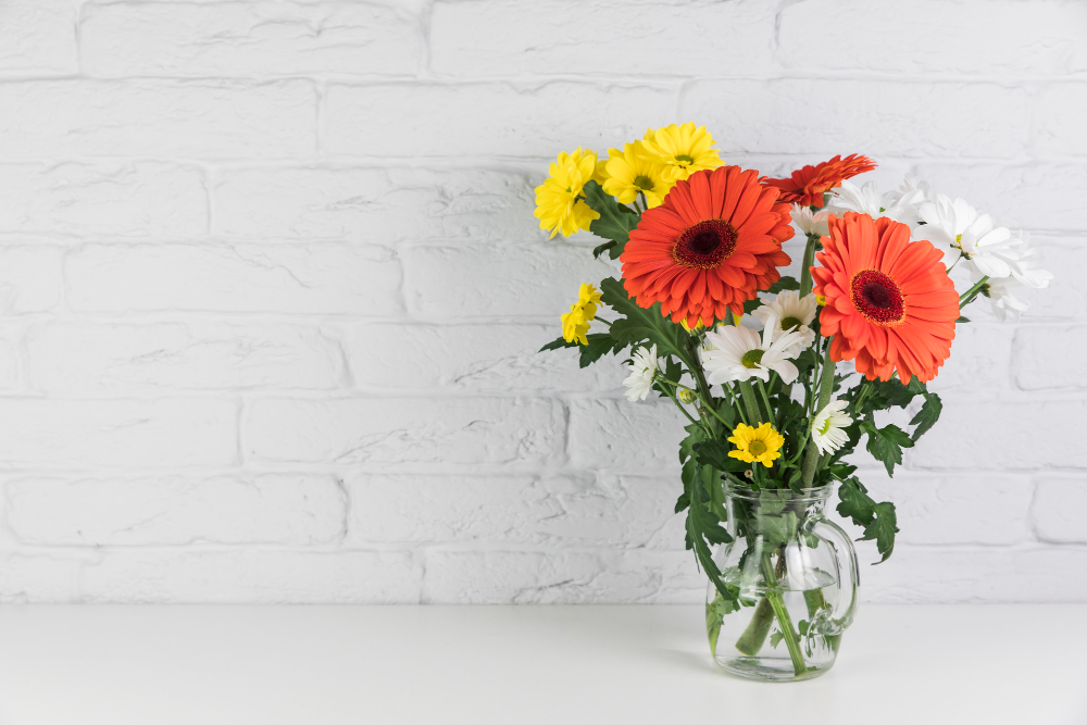 chamomile-gerbera-flowers-glass-jug-desk-against-white-brick-wall